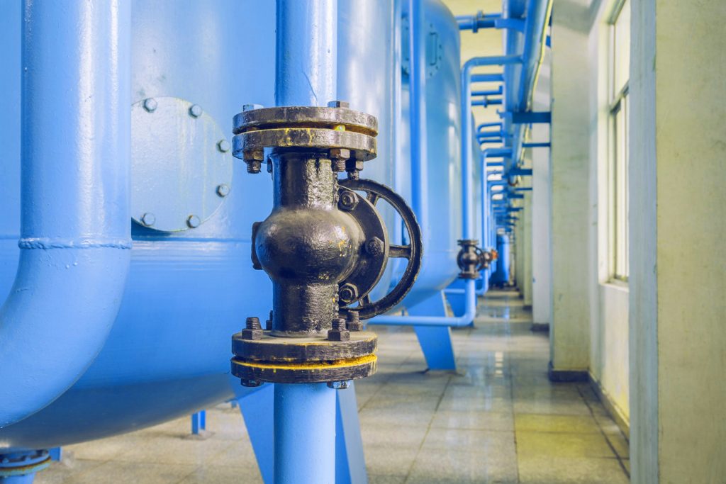 Schwarze Rohrleitung mit einem Absperrventil, Teil eines Wärmetauschersystems. Im Hintergrund sind weitere blaue Rohre und Druckbehälter in einer industriellen Umgebung zu sehen.